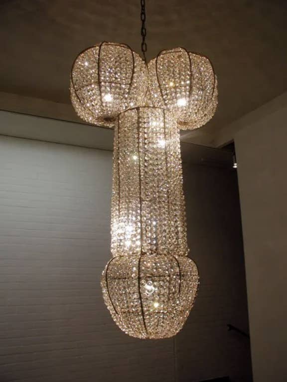 An ornate chandelier with multiple lighted sections hangs from a ceiling, looking like a penis