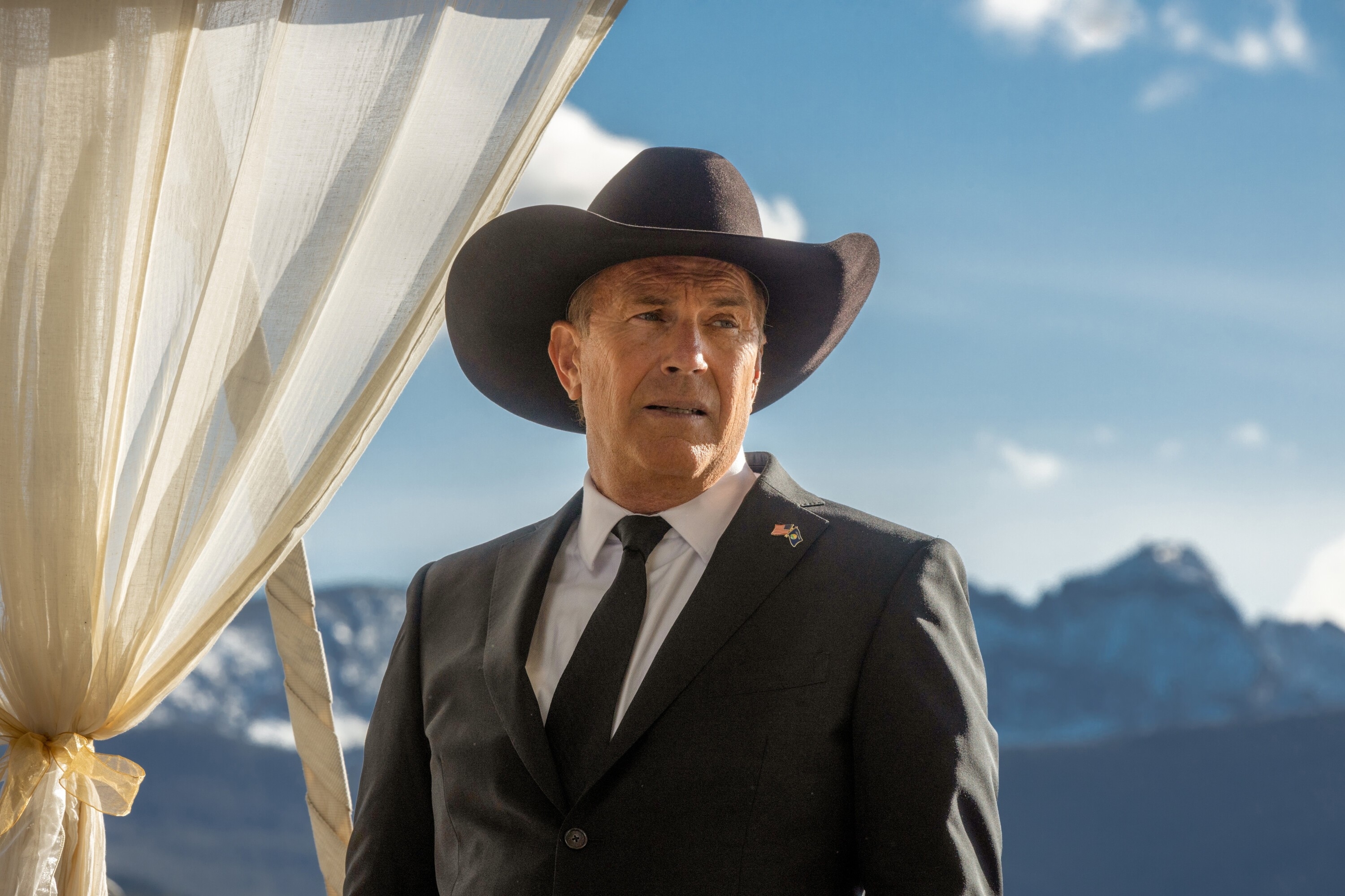 Kevin Costner in character as John Dutton from &quot;Yellowstone,&quot; wearing a suit and cowboy hat with mountains in the background