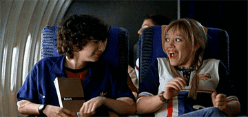 Dos personajes animados están en un avión; el personaje masculino a la izquierda sostiene un libro mientras la femenina a la derecha se ríe