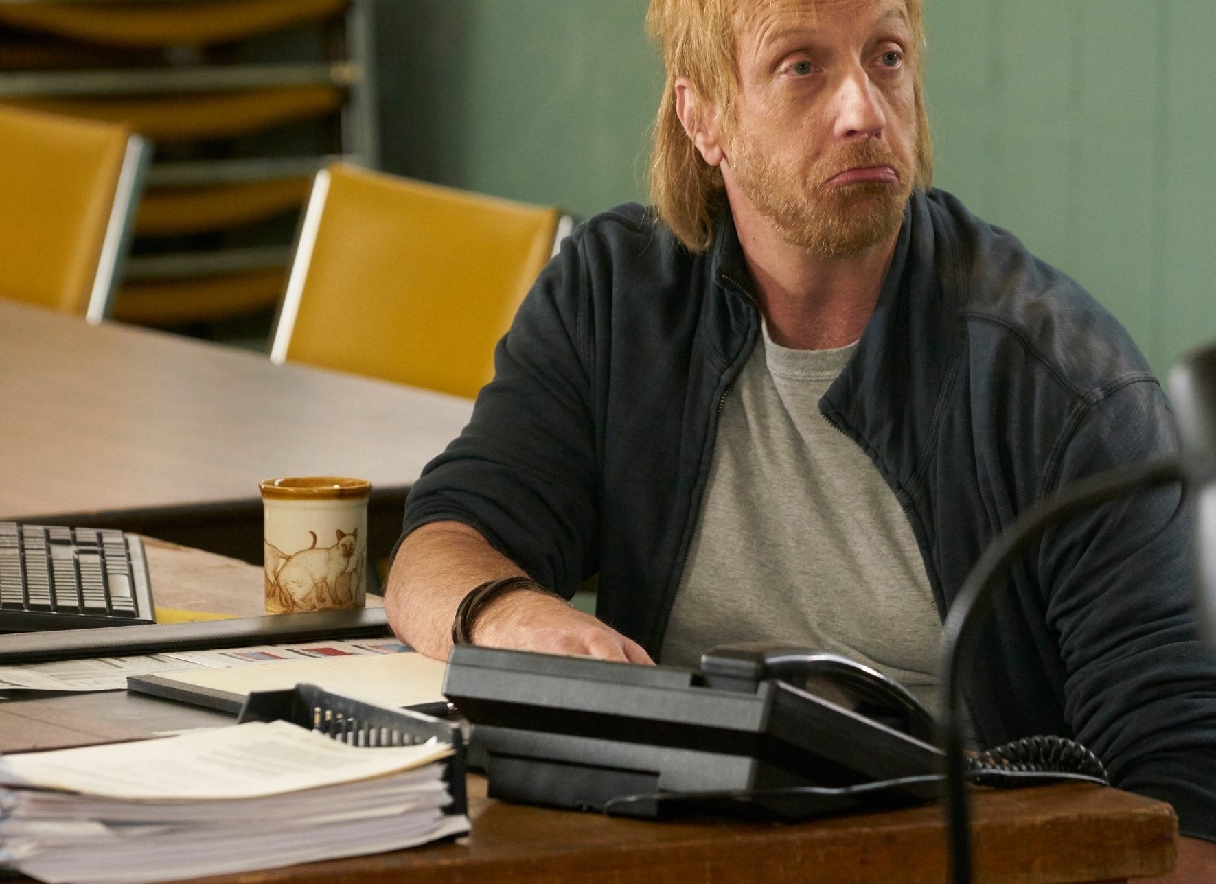 Man sitting at a desk with phones, looking pensive in a TV show scene
