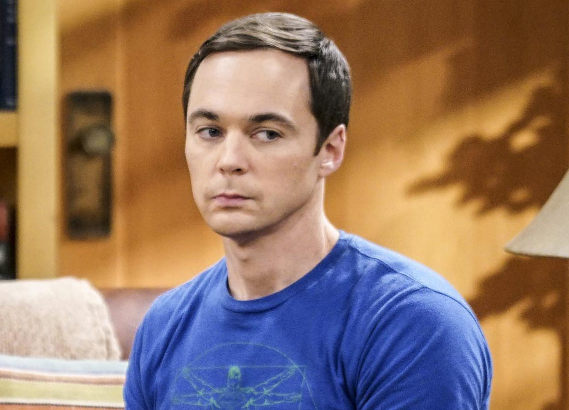 Sheldon Cooper from Big Bang Theory wearing a blue t-shirt with a graphic, seated with a serious expression