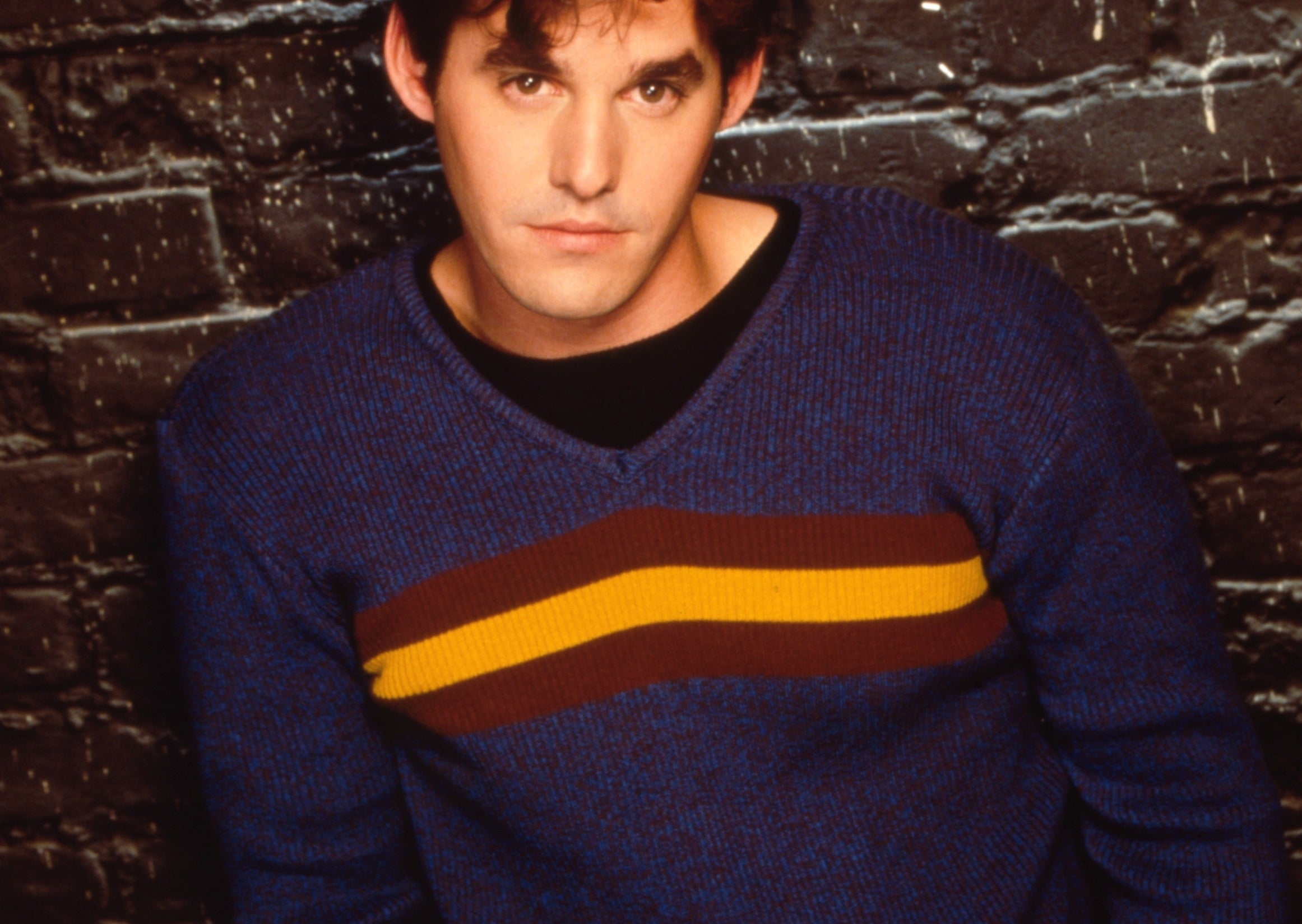 Man in a blue sweater with a yellow and red stripe, leaning against a brick wall