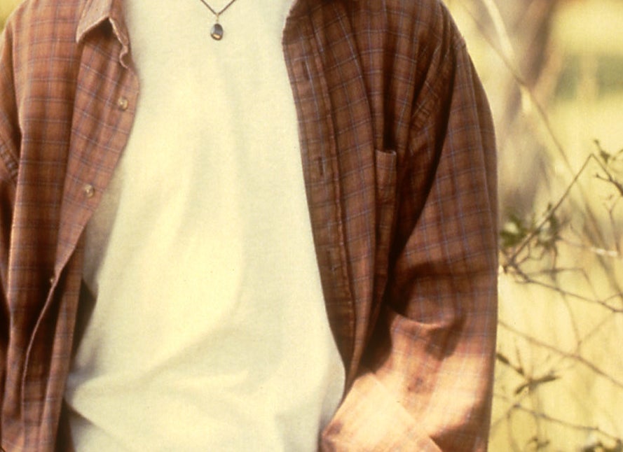 Man standing in a field, wearing an open flannel shirt over a t-shirt, with a casual, relaxed pose