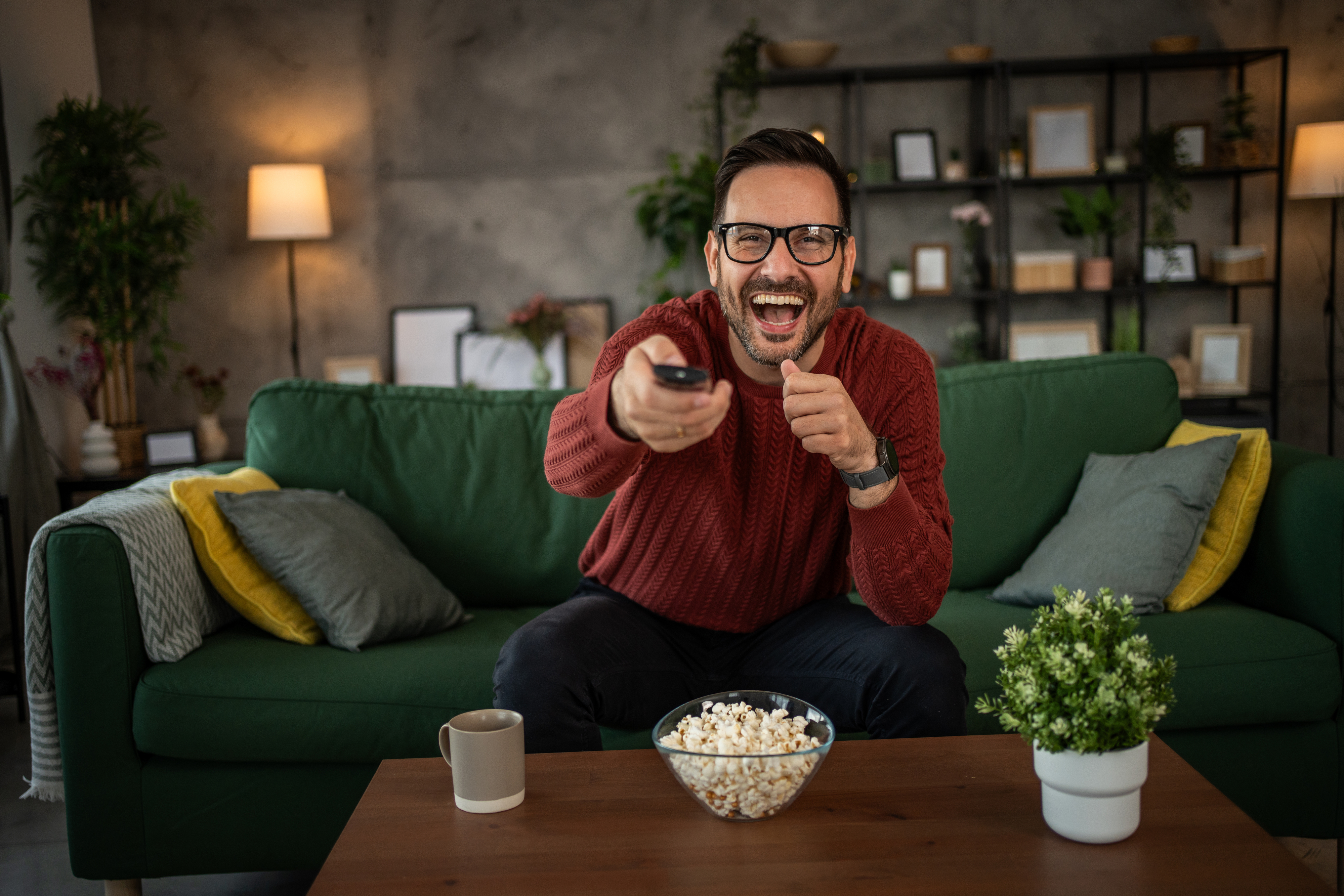 Man in sweater laughing and pointing remote at TV in a living room with a couch, popcorn, and coffee mug