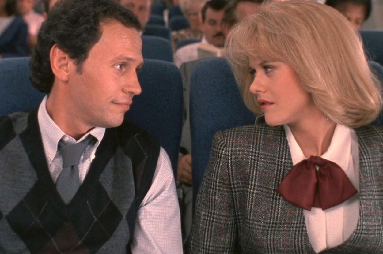 Two characters from a film sitting side by side on an airplane; man in a sweater vest, woman in a suit with a bow tie