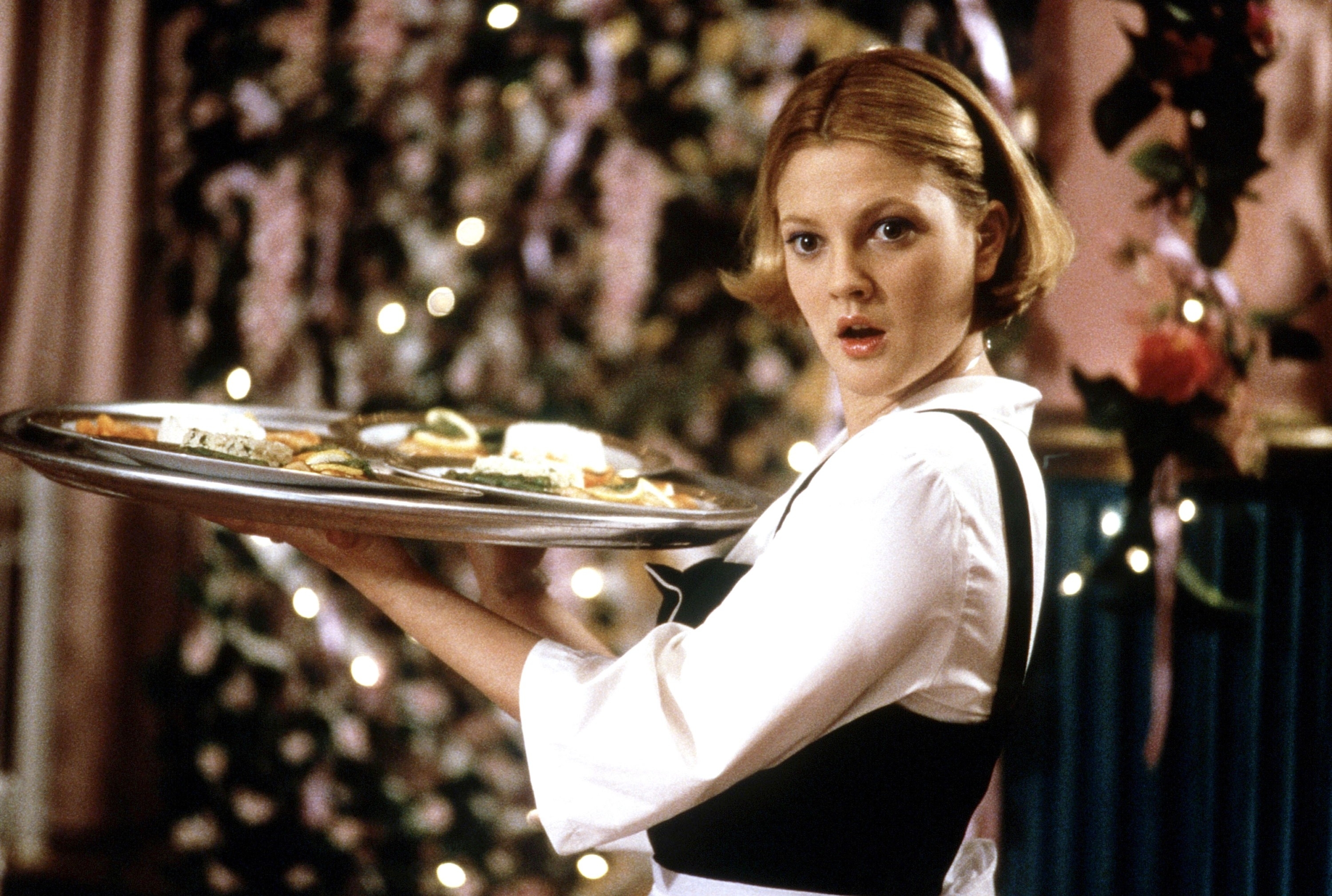 Drew Barrymore in a server uuniform holding a tray with food, standing in front of flowers and greenery