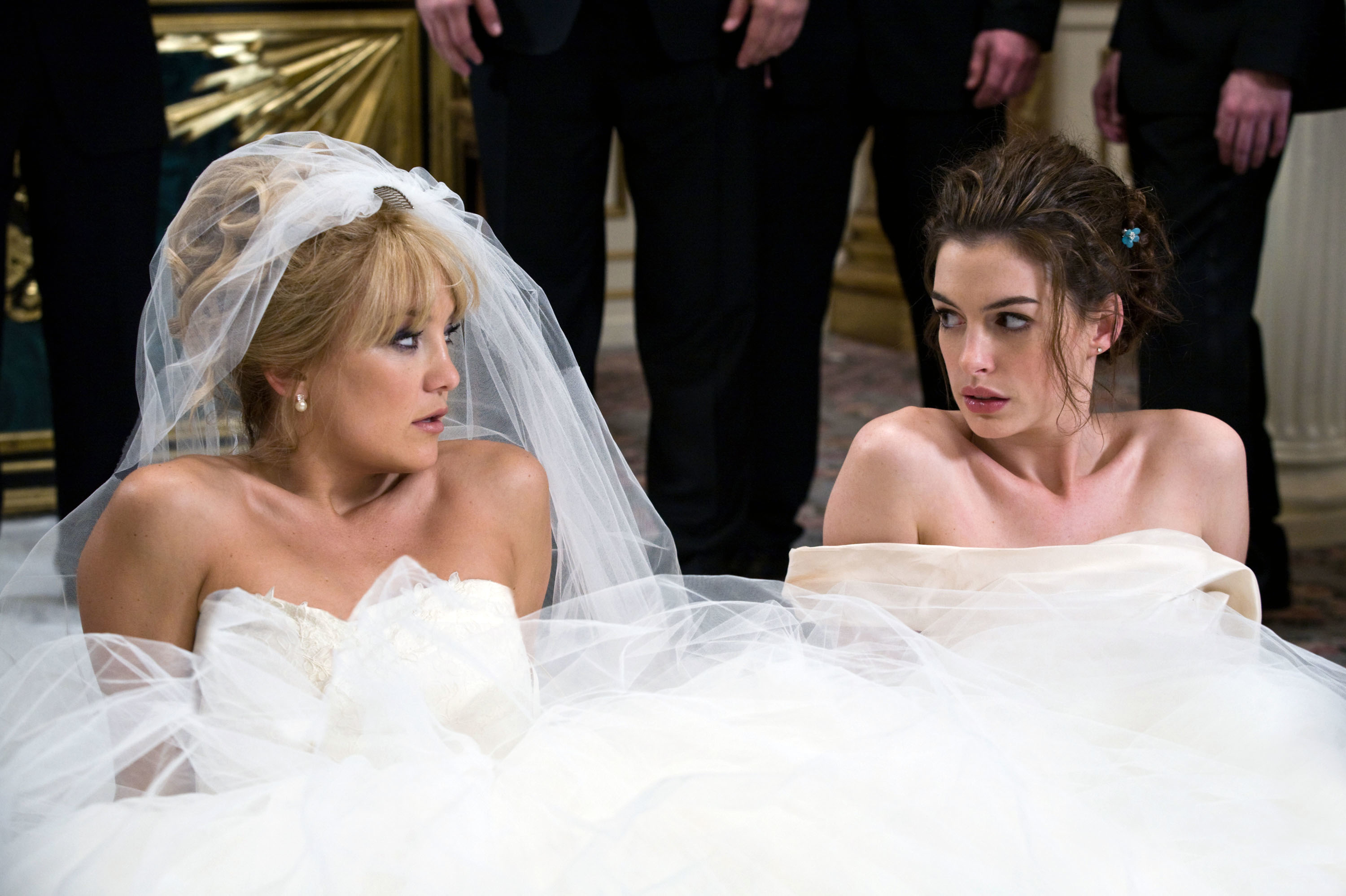 Kate Hudson and Anne Hathaway seated side by side look at each other, one in a veil and the other bare-shouldered