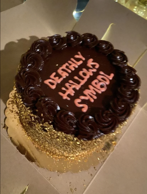 Chocolate cake with "Deathly Hallows Symbol" written in icing, topped with gold sprinkles