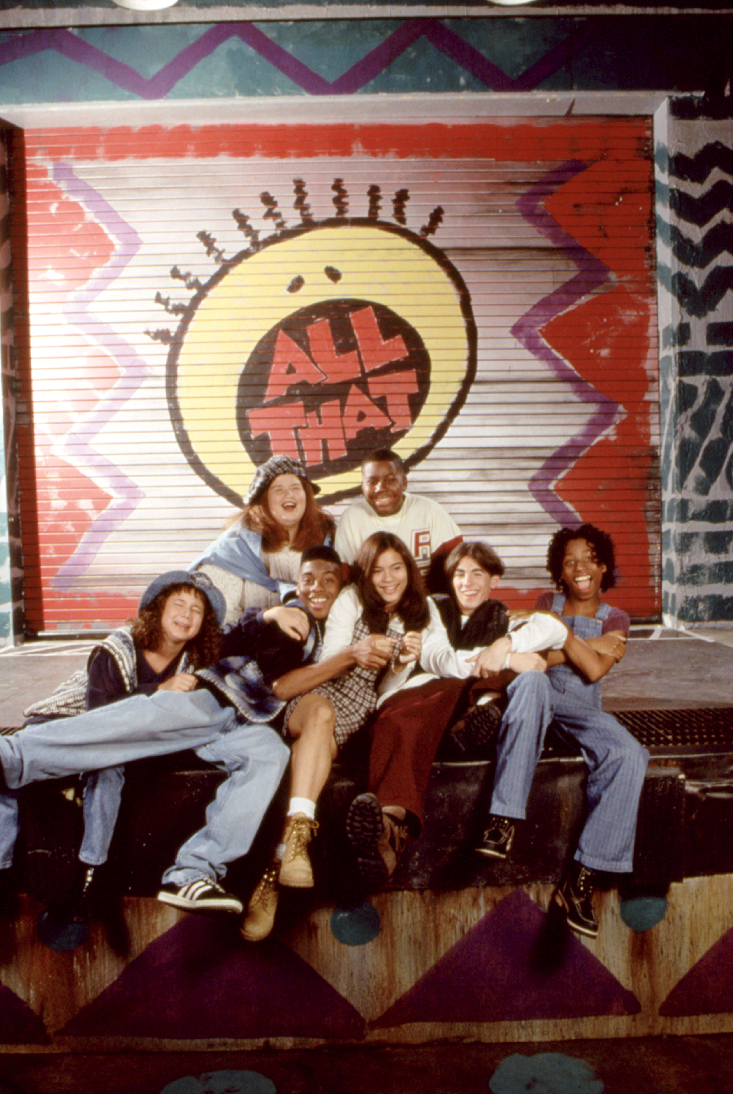 Cast of &quot;All That&quot; posing in front of show&#x27;s logo, in casual 90s attire, smiling