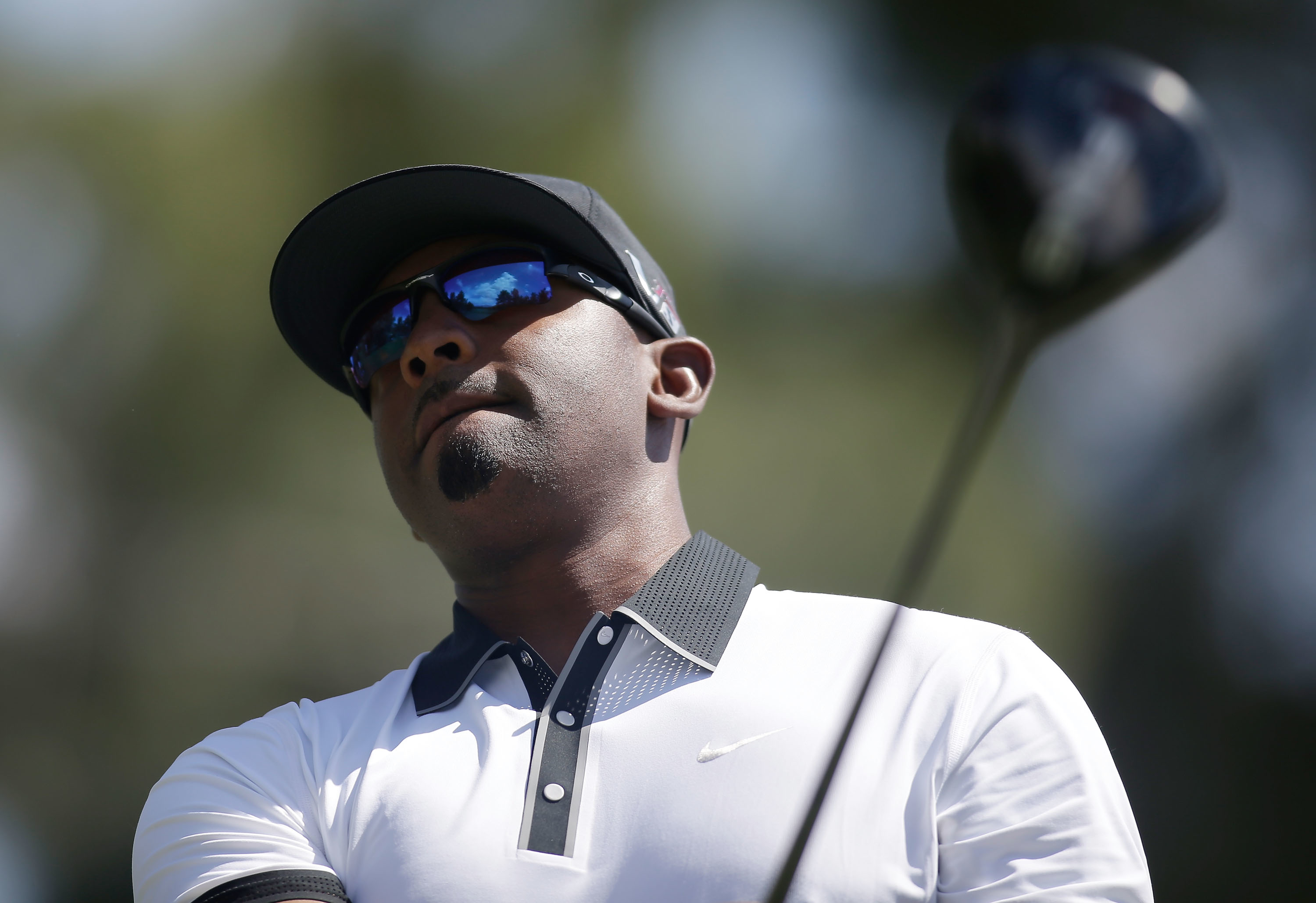 Golfer mid-swing with club, focused expression, wearing a cap, sunglasses, and sporty attire