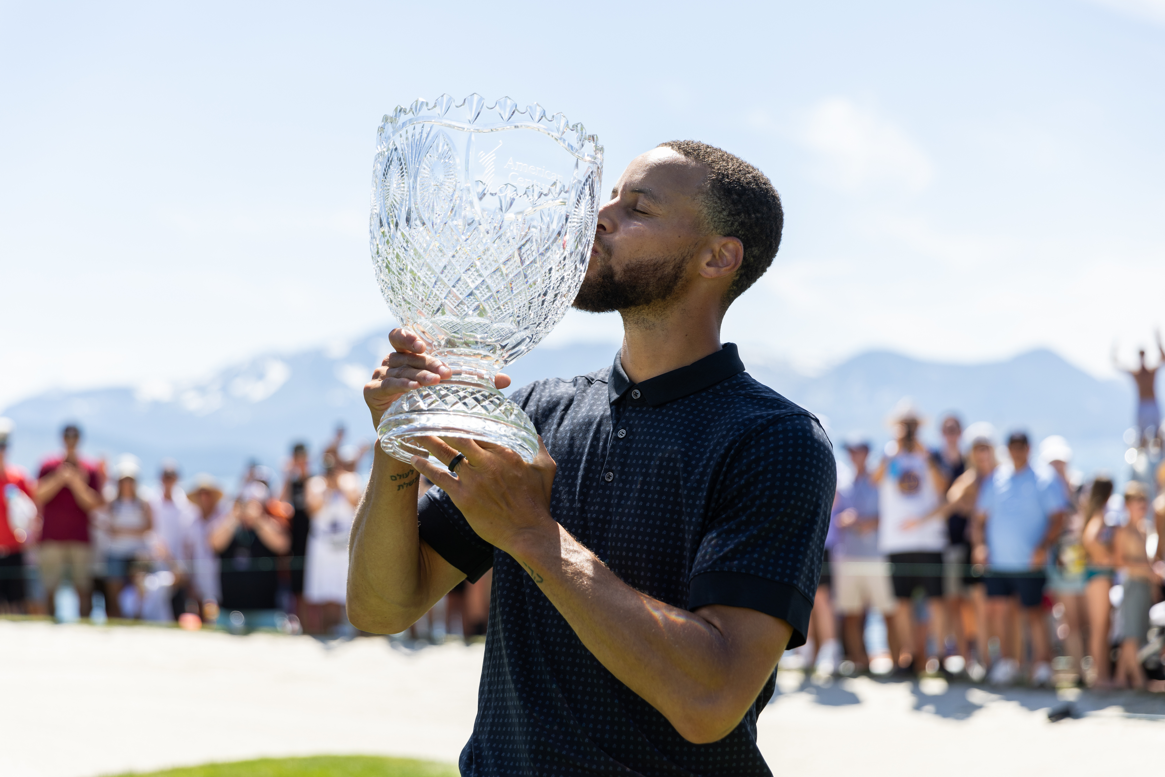 Athlete in polo shirt kisses trophy at golf course with spectators and mountains in the background