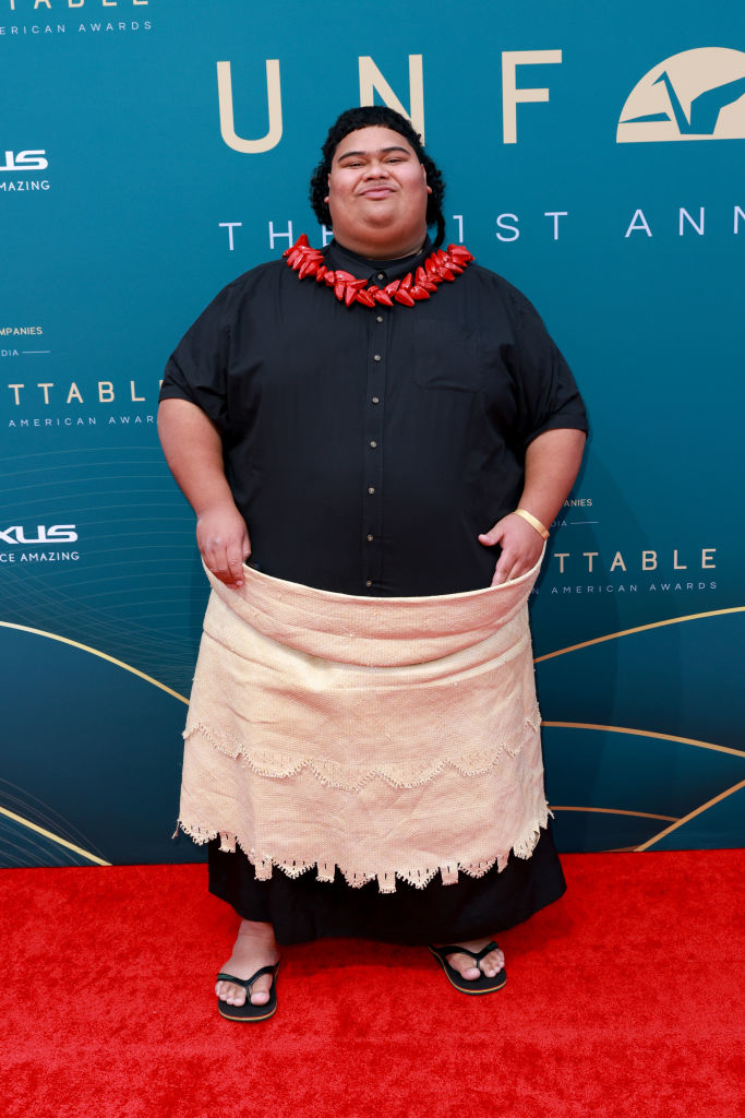 Individual wearing a black shirt, tan lavalava skirt, and sandals on a red carpet