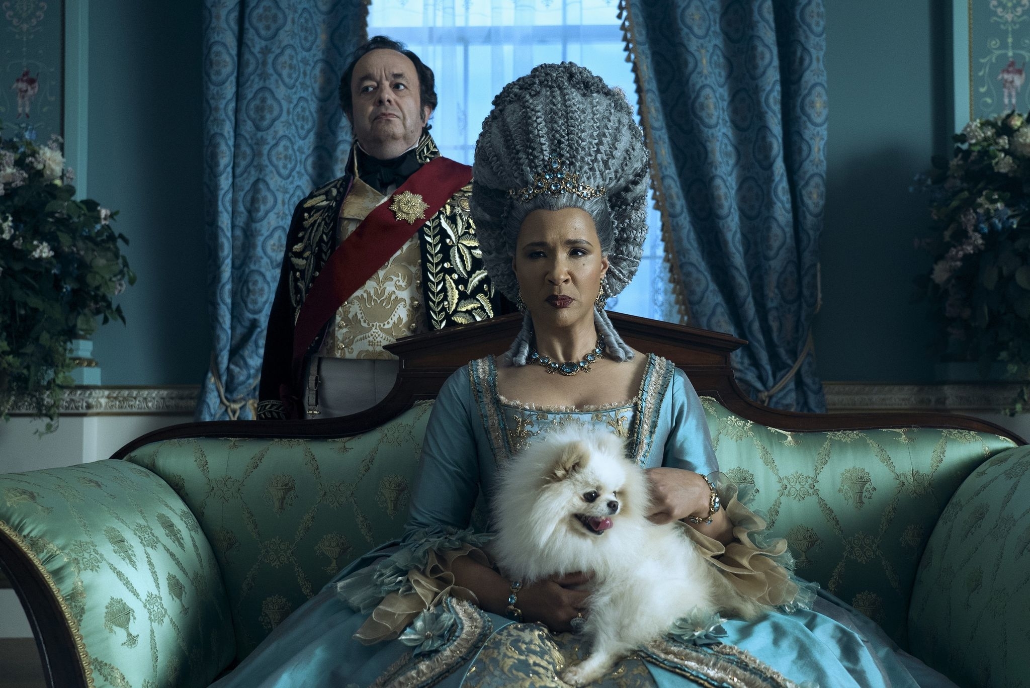 Dos personajes de época sentados, mujer con vestido azul y hombre con traje rojo, junto a un perro blanco