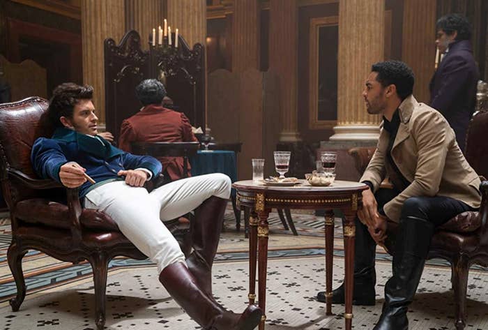 Personajes de una serie sentados en una elegante sala, con trajes de estilo moderno. Dialogan en un ambiente de tensión