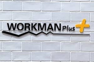 Sign reading "WORKMAN Plus+" on a white brick wall above an office equipment stand