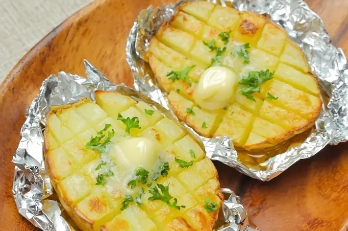 フォイルに包まれた焼きジャガイモ2つが木製の皿に乗せられている。上にバターとパセリが添えられている。