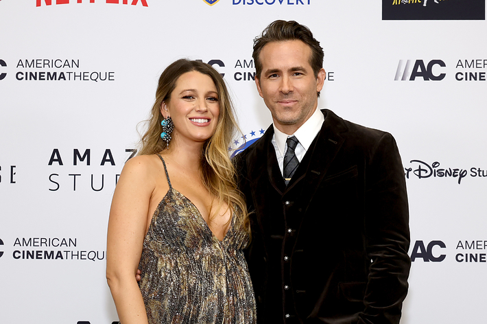 Blake Lively in a patterned dress with Ryan Reynolds in a suit, both smiling on a sponsor backdrop