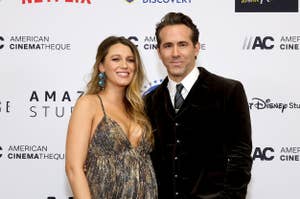 Blake Lively in a patterned dress with Ryan Reynolds in a suit, both smiling on a sponsor backdrop