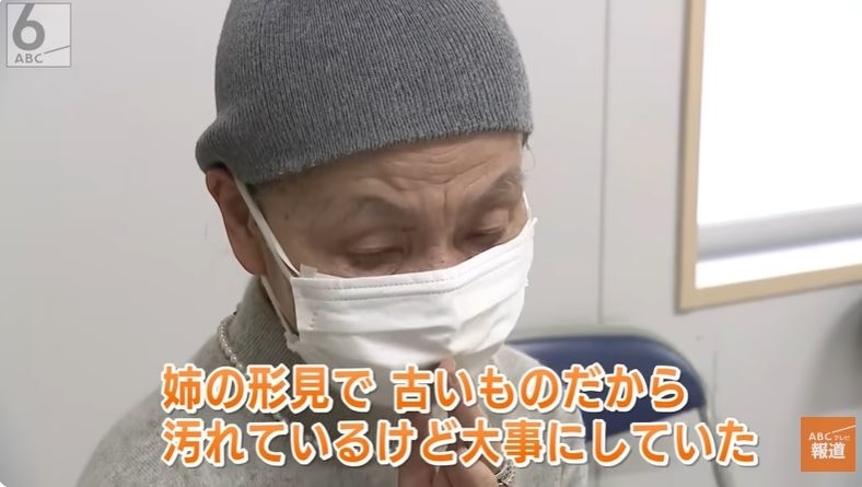 この画像は、マスクをした中年の男性が目を閉じて頭を抱える様子を映しています。画面下部には日本語のテキストがあります。