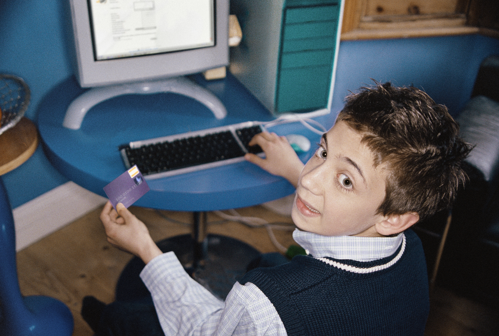 Young person at computer desk, holding up a card, looking back with a surprised expression