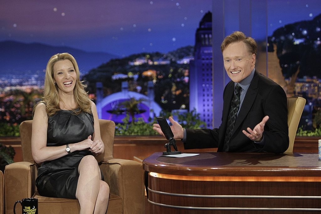 Man in a suit and woman in a black dress sitting, talking on a talk show set