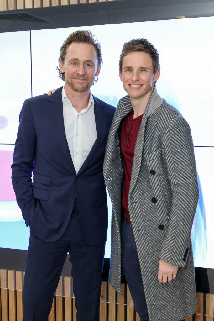 Two men standing side by side, one in a blue suit and white shirt, the other in a grey coat and red scarf