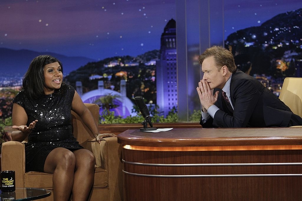 Two people, one reclining on a couch, another seated at a desk, engage in conversation on a talk show set