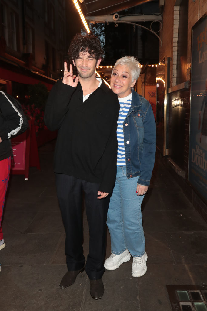 Two people smiling and posing together on a street, one making a peace sign