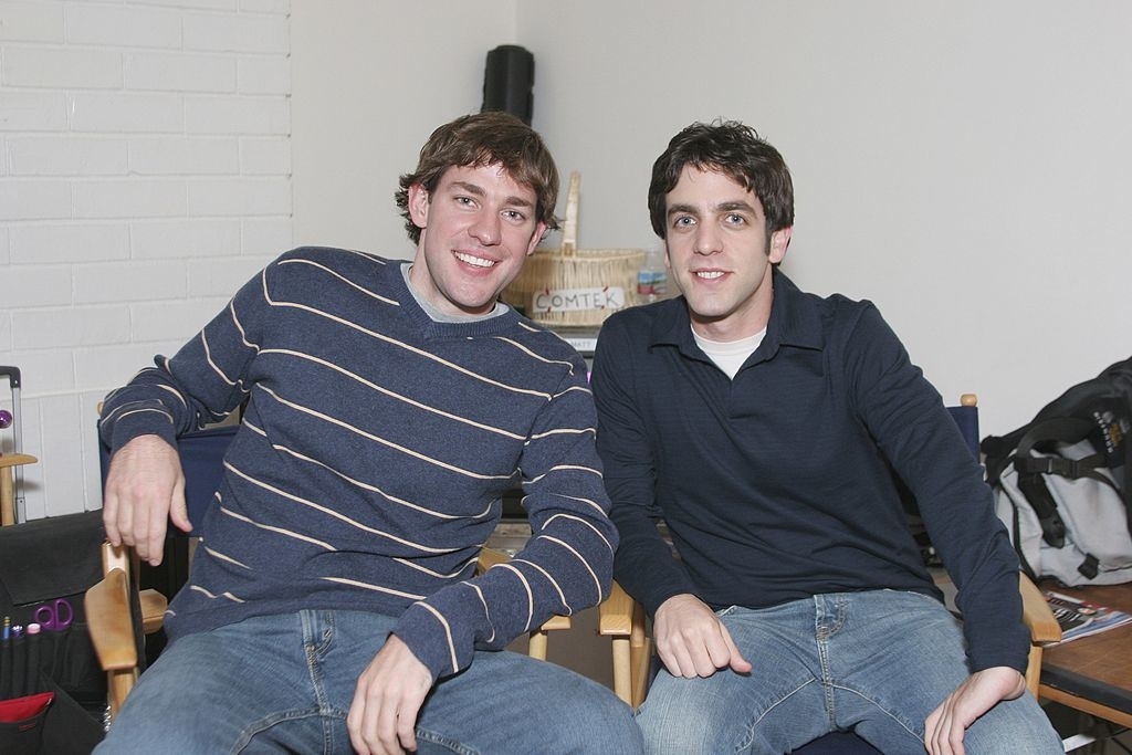 Two men smiling and seated next to each other, casually dressed in jeans and long-sleeve shirts