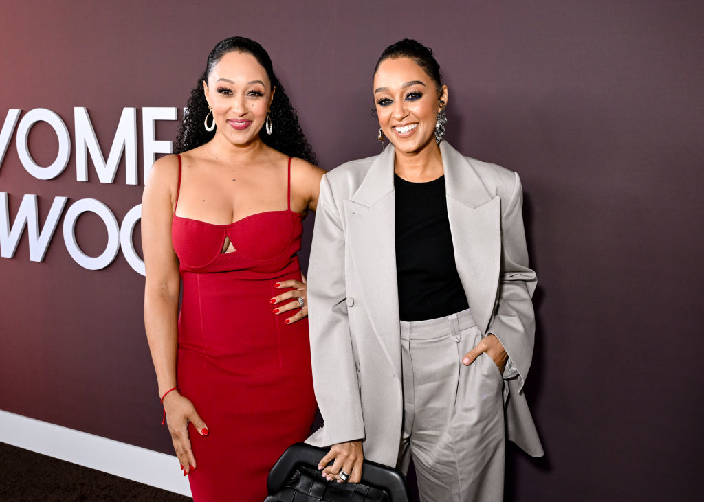 Two women posing together; one in a red sleeveless dress, the other in a grey suit with a black top. Both are smiling
