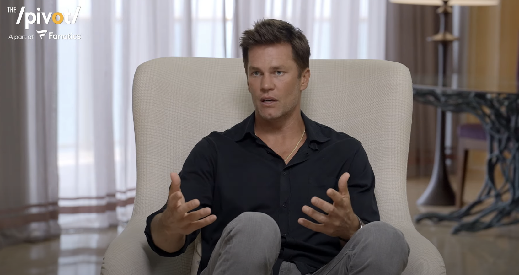 Tom Brady in a casual black shirt, gesturing while sitting and speaking during an interview