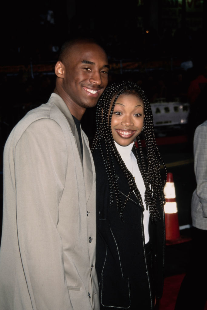 Kobe Bryant and Brandy smile at an event. He wears a suit; she has braids and a chic outfit
