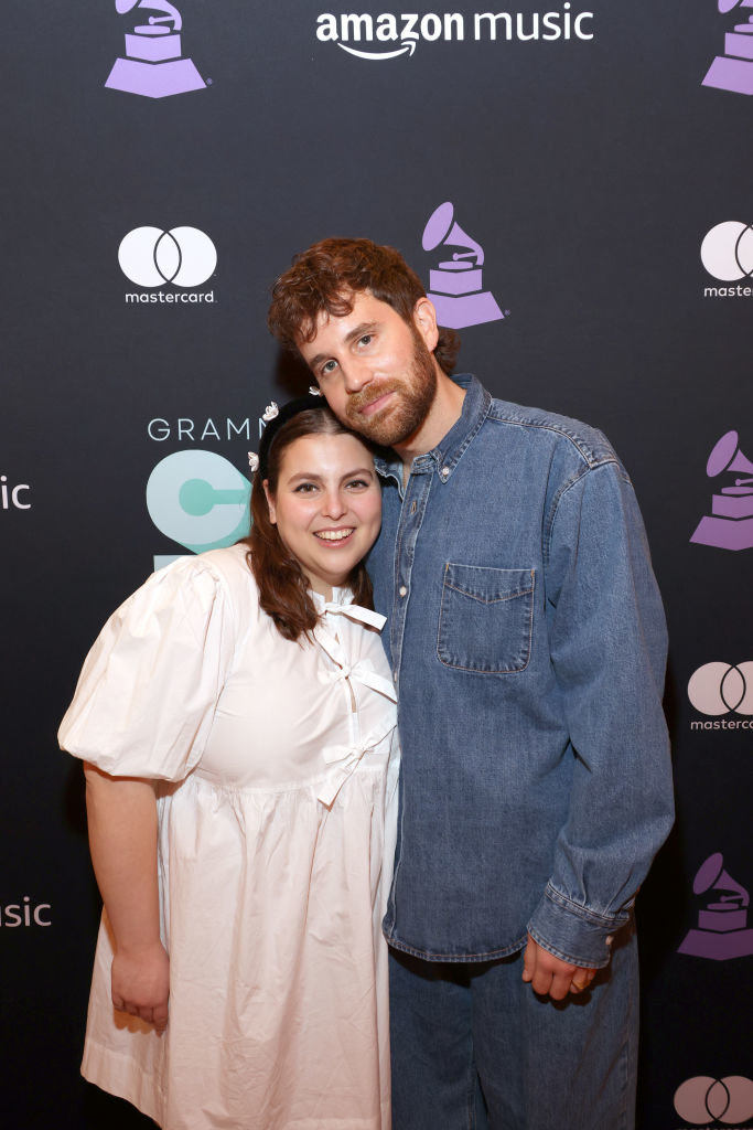 Two individuals posing together; one in a white puff-sleeve dress, the other in a denim outfit, at an event with branded backdrops