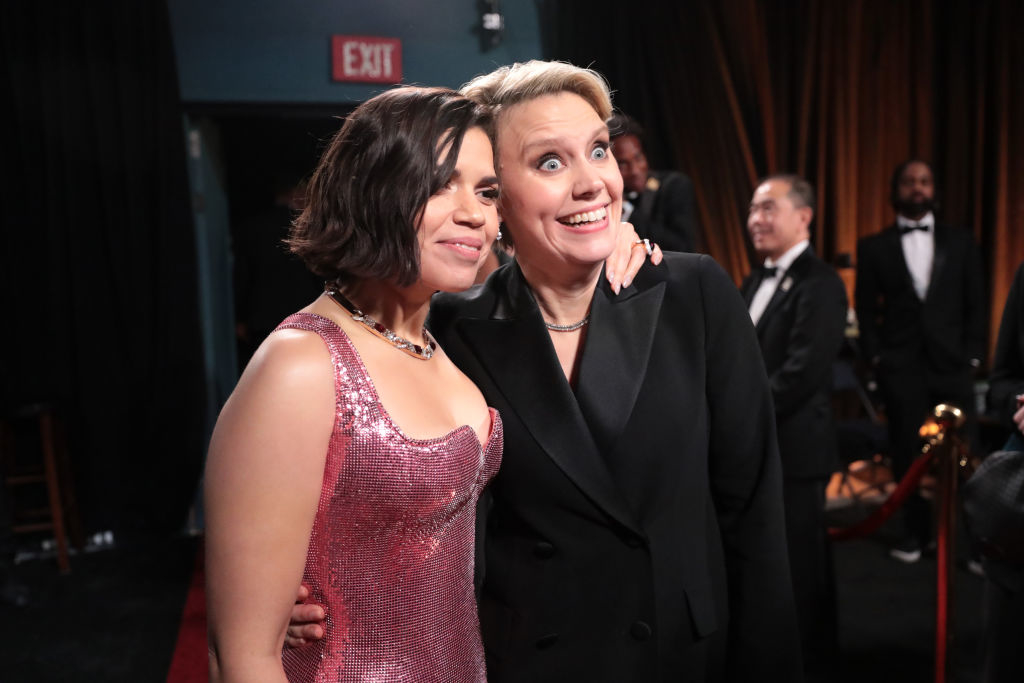 Two women smiling at an event, one in a glittery gown, the other in a black suit