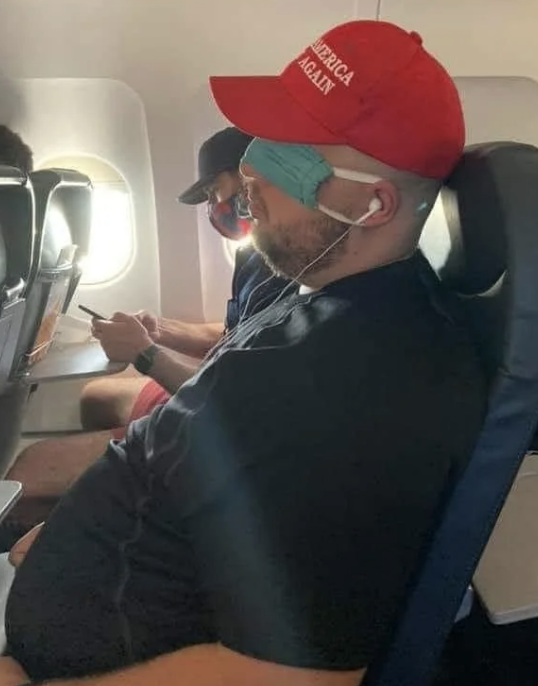 Person in airplane wearing a cap with text, mask under chin, others seated nearby
