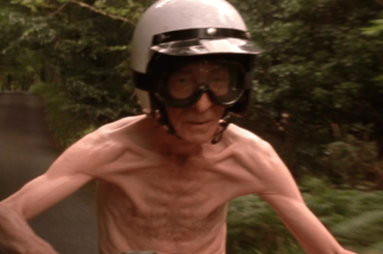 Elderly gentleman wearing a helmet and goggles, riding a motorcycle with trees in the background