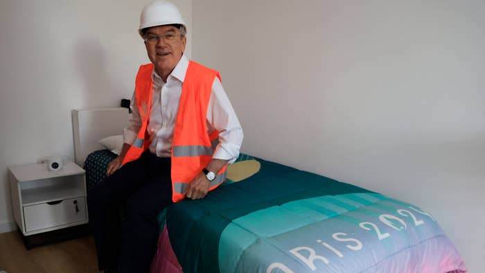 Man in hard hat and hi-vis vest sits on bed with &quot;Paris 2024&quot; logo, in Olympic Village room