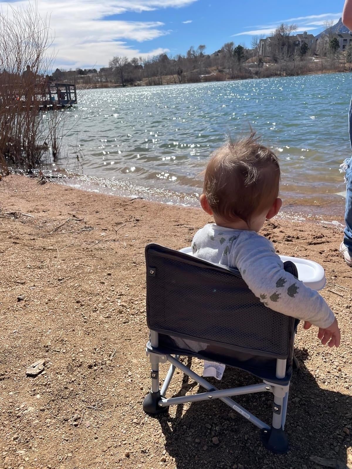 Toddler successful  a portable seat  facing a water  with a dock successful  the background, with an adult's limb  beside the chair