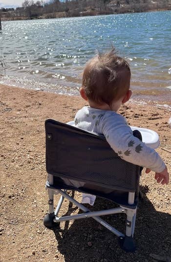 Toddler successful a portable seat facing a water with a dock successful the background, with an adult's limb beside the chair