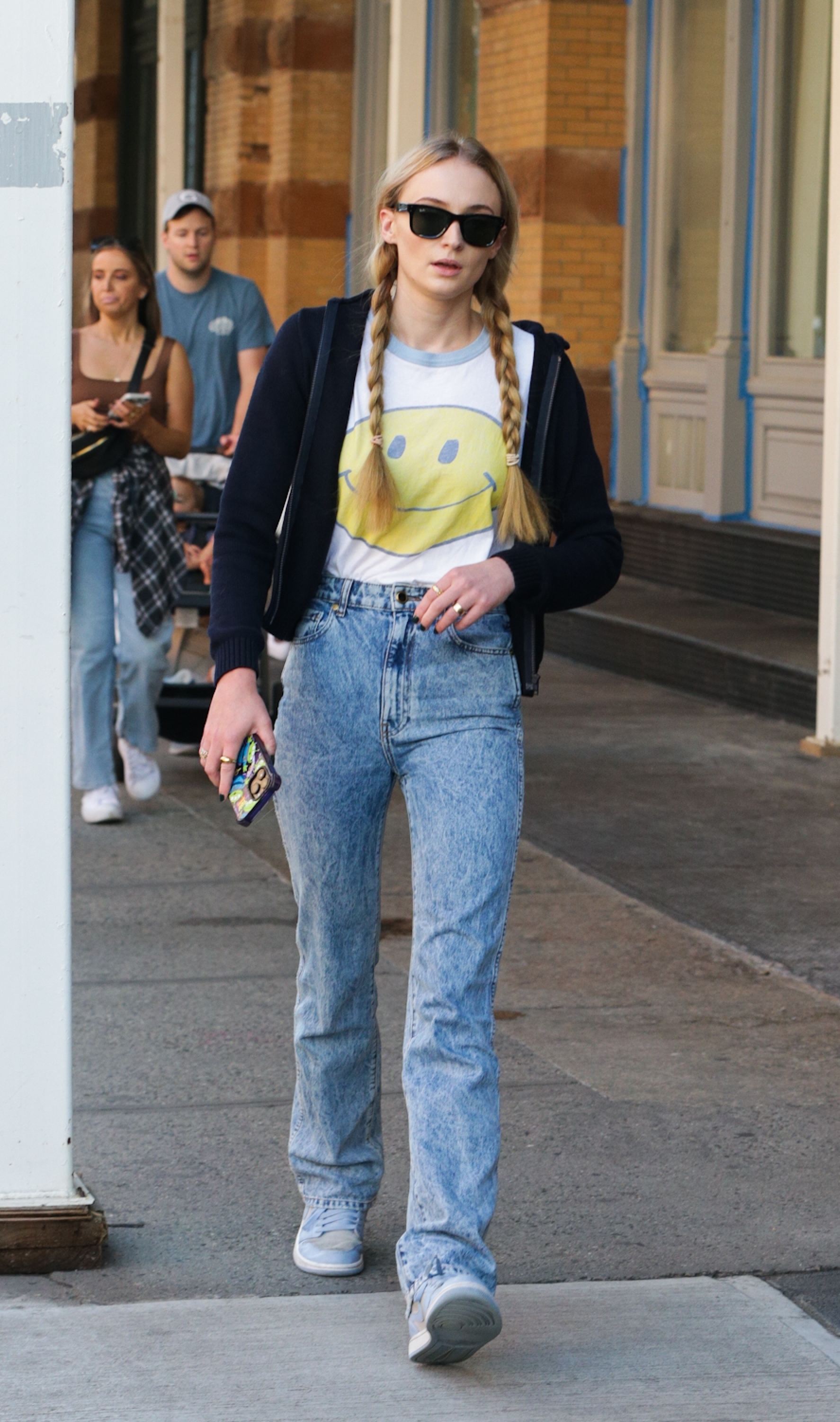 Sophie Turner in casual attire with braided hair, graphic tee, jeans, and sneakers walks on a sidewalk