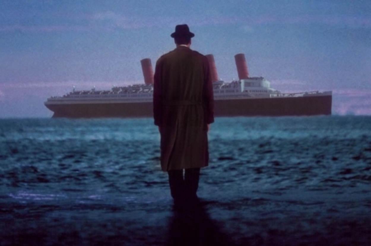 Man in a long coat looking at a large ship in the distance at sea