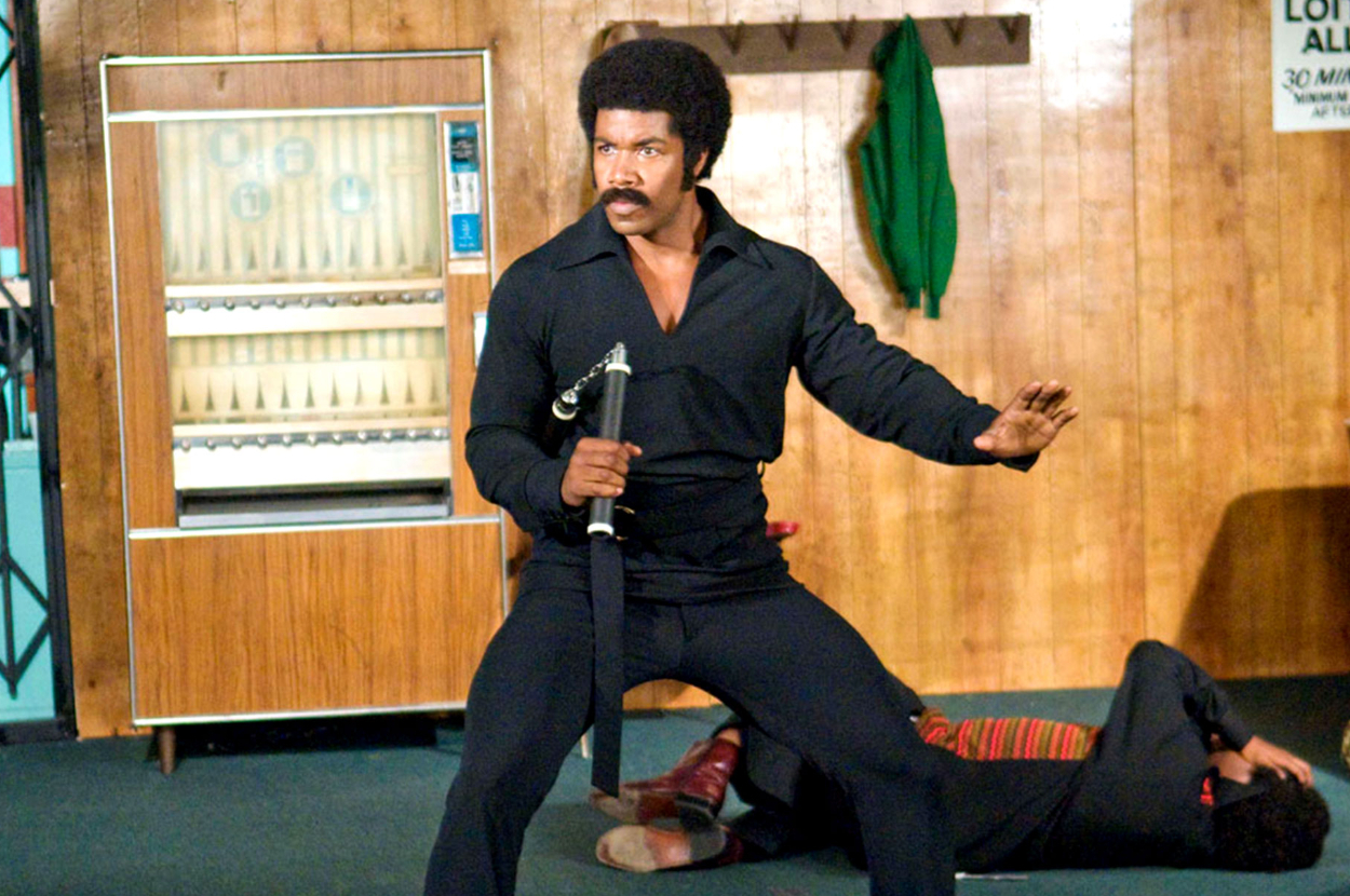 Character Black Dynamite in an action pose with a nunchaku, another character lies on the ground