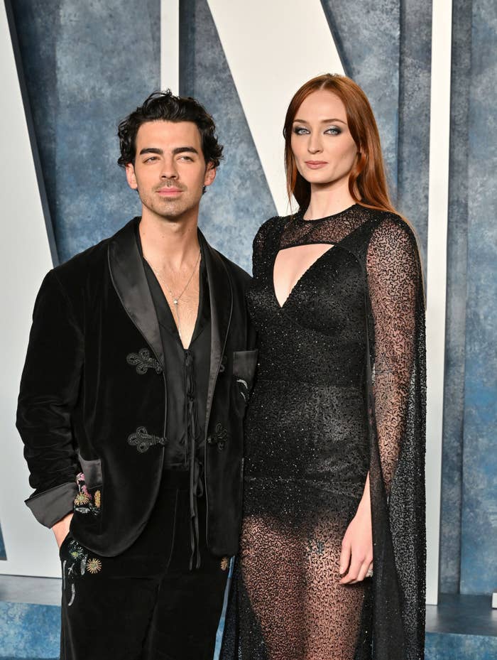 Joe Jonas and Sophie Turner stand side by side; one in a dark embellished velvet jacket, the other in a sparkling black cutout dress