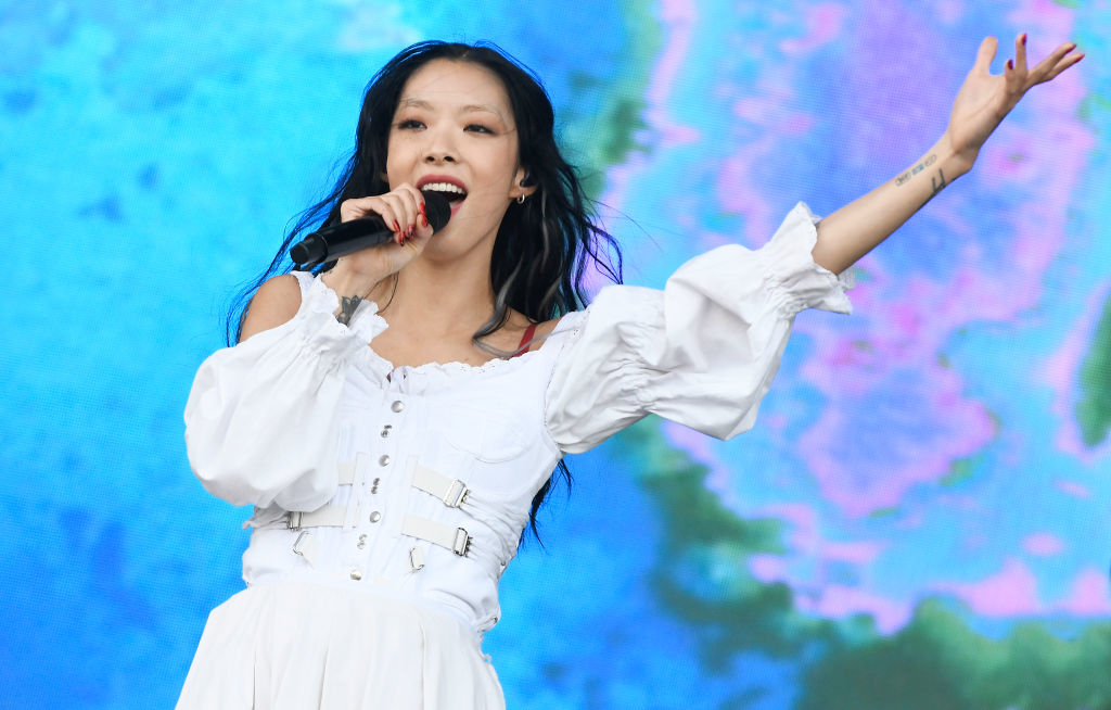 Rina in a longsleeved dress singing on stage with a microphone, arm raised