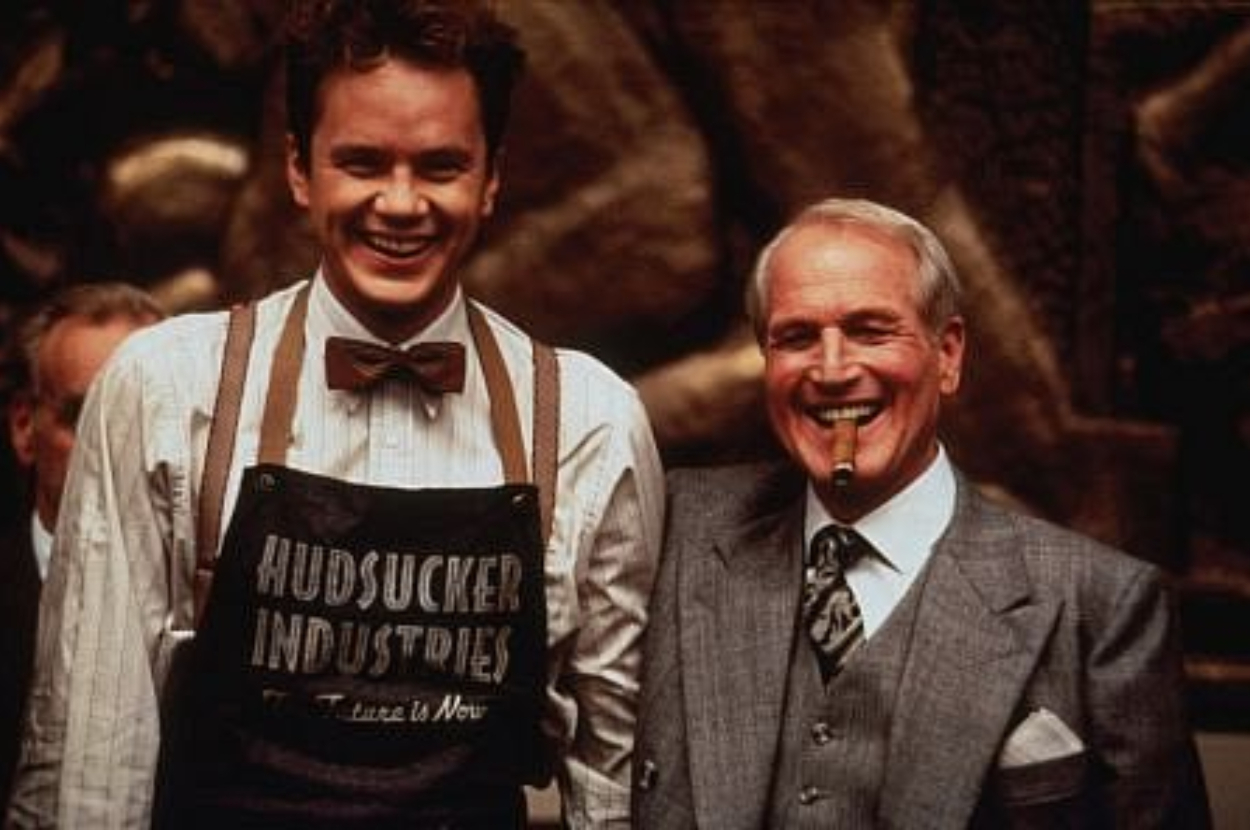 Two characters smiling, one wearing a &quot;Hudsucker Industries&quot; apron and bowtie, the other in a suit