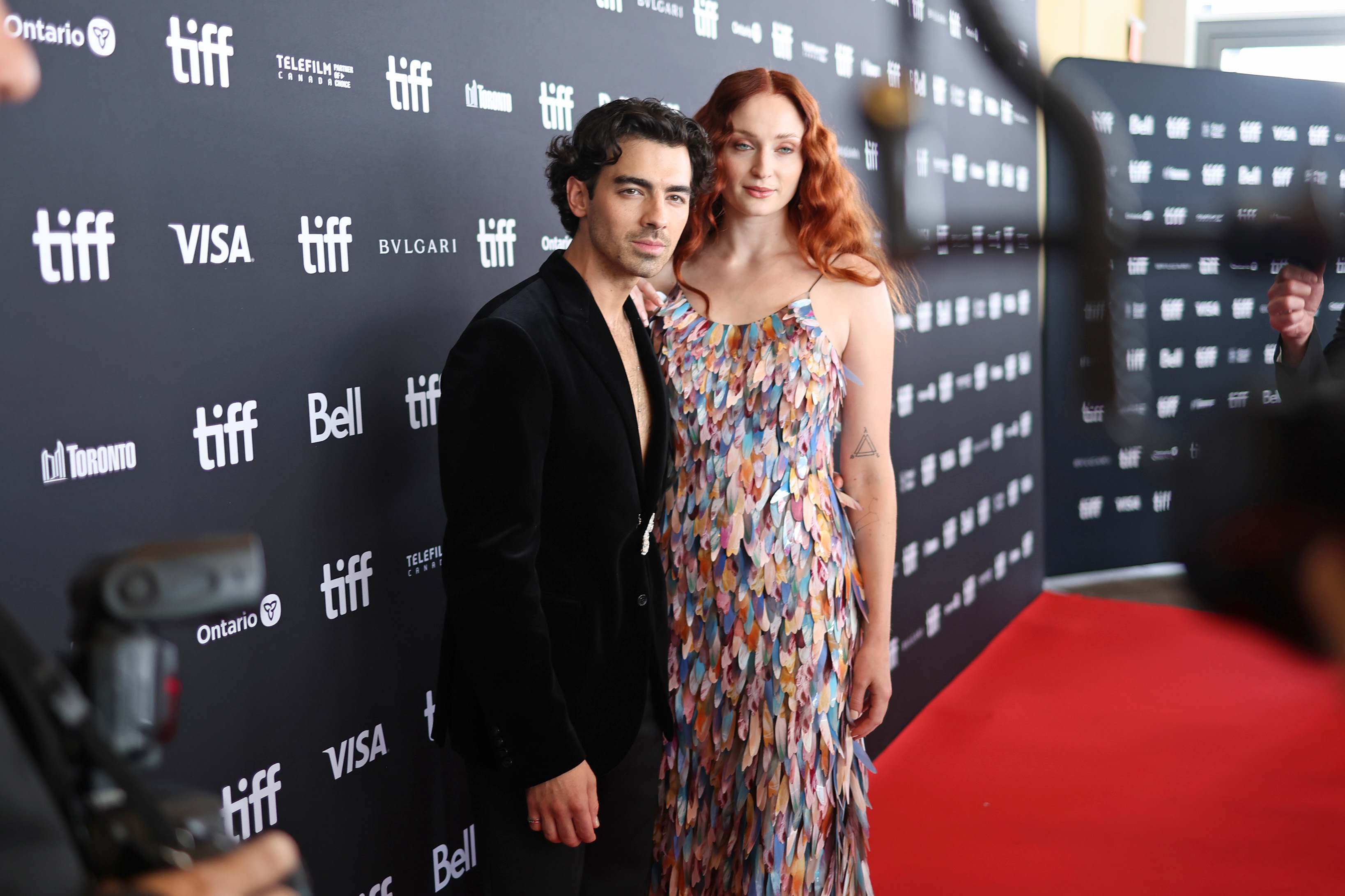 Joe Jonas and Sophie Turner pose together at a TIFF event; the man in a black suit and the woman in a floral dress