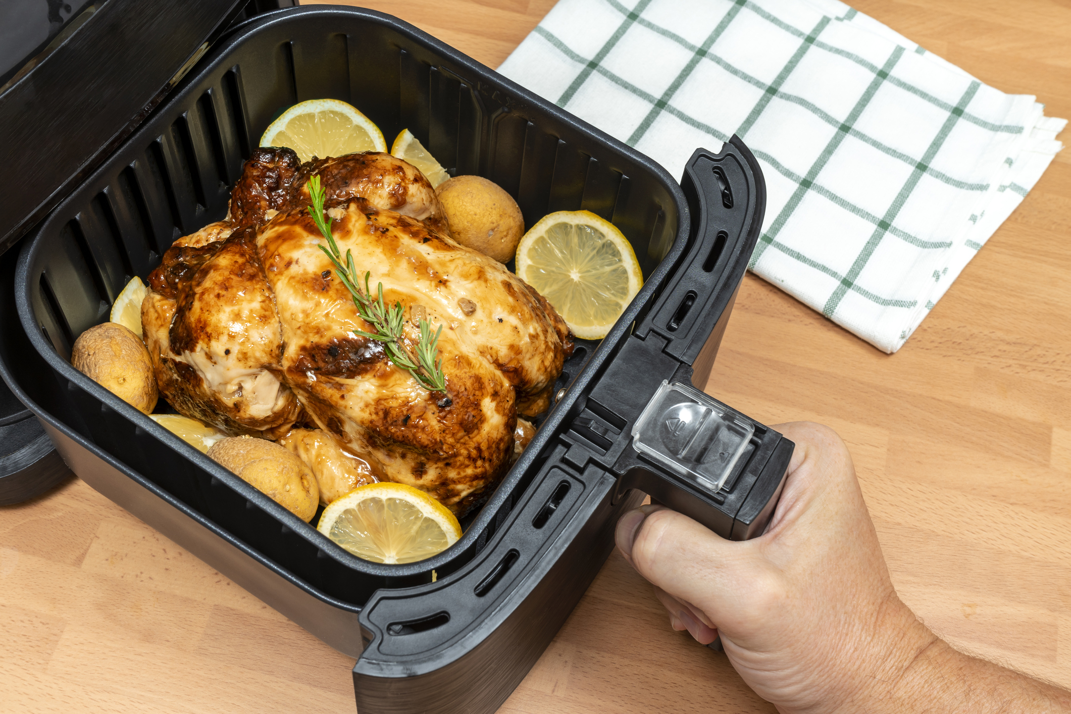 A person&#x27;s hand lifting the lid of an air fryer containing a roasted chicken and potatoes