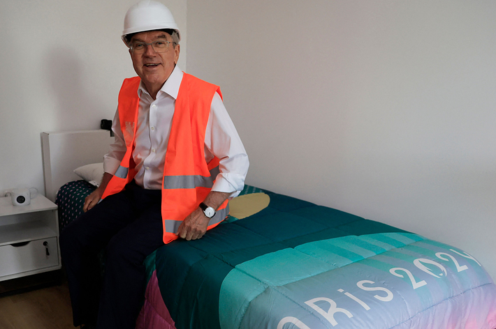 Man in hard hat and hi-vis vest sits on bed with "Paris 2024" logo, in Olympic Village room