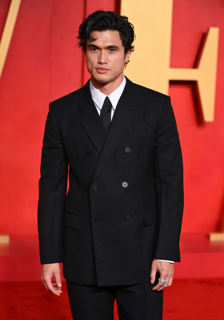 Charles Melton in a double-breasted suit stands confidently at an event