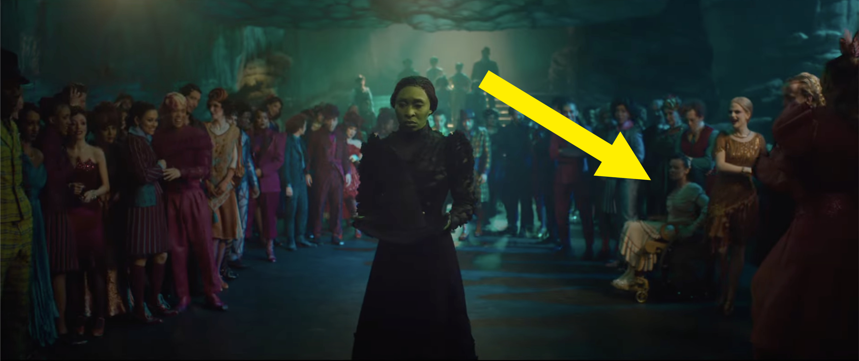 A still from a film showcasing a character in a mask standing center with onlookers in costume surrounding them